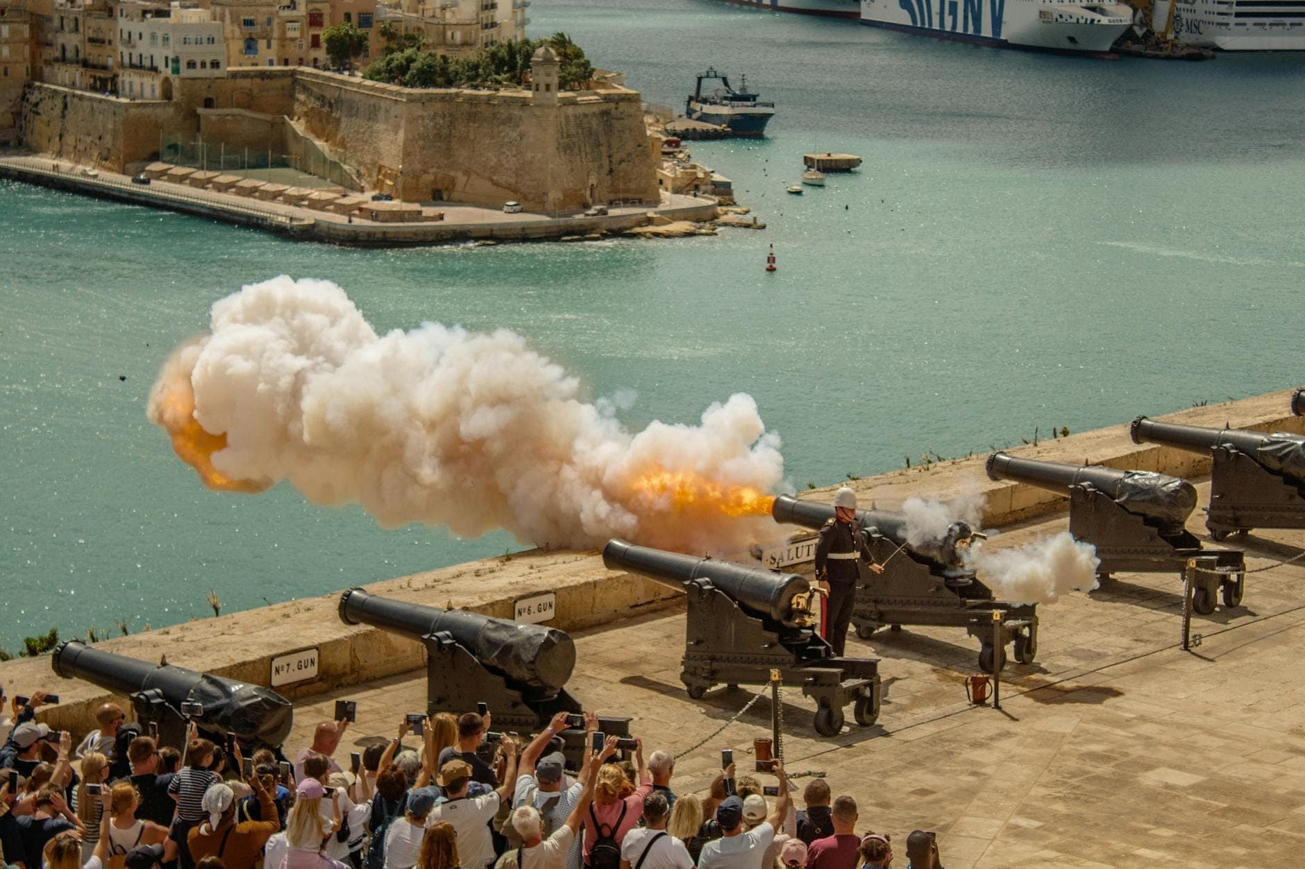 Crowds watch the noon cannon fire and smoke erupting from the historic Saluting Battery at Upper Barrakka Gardens, overlooking Malta’s Grand Harbour.